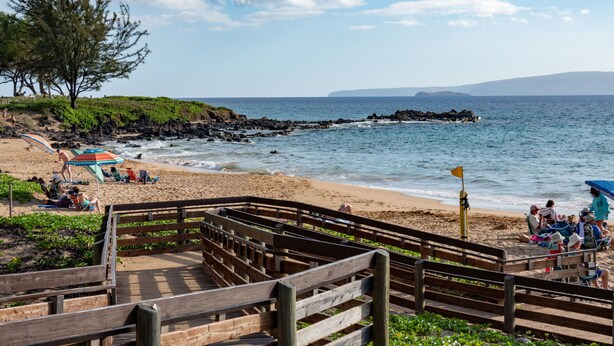 A wooden boardwalk leading to a beach with people relaxing under umbrellas. - KIHEI AKAHI CONDO STEPS FROM BEACH/GARDEN & PEEKABOO OCEAN VIEW/POOL-HOTEL ZONED (Kihei)