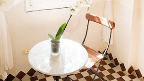 A room with a checkered floor, a round table with a potted plant, a chair, and sheer curtains over a window. - Il Rifugio Pecorella (Alvignano)