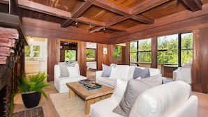 A spacious living room with a high ceiling, wooden beams, white sofas, and large windows offering a view of greenery.