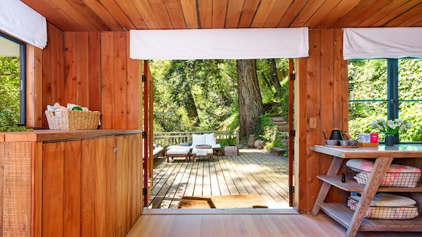 A wooden cabin interior with a deck leading to a forested area, a small table with items, and a wicker basket on a shelf.