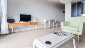 Una sala de estar moderna con un televisor de pantalla plana, una mesa de centro blanca con revistas, un sillón verde y una mesita blanca con un florero.