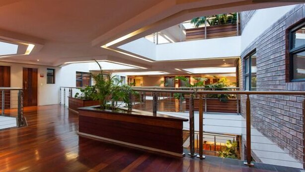 Un vestíbulo amplio con suelo de madera, una jardinera central con plantas y una barandilla de cristal que da a un nivel inferior.