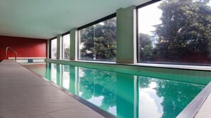 Una piscina con agua cristalina, rodeada de amplios ventanales que ofrecen vistas a árboles y edificios.