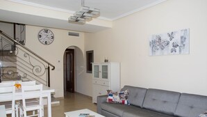 Ein Wohnzimmer mit grauem Sofa, weißem Couchtisch und einem Essbereich mit weißem Tisch und Stühlen. Eine Treppe mit einer Uhr an der Wand und ein Schrank mit einem Fernseher.
