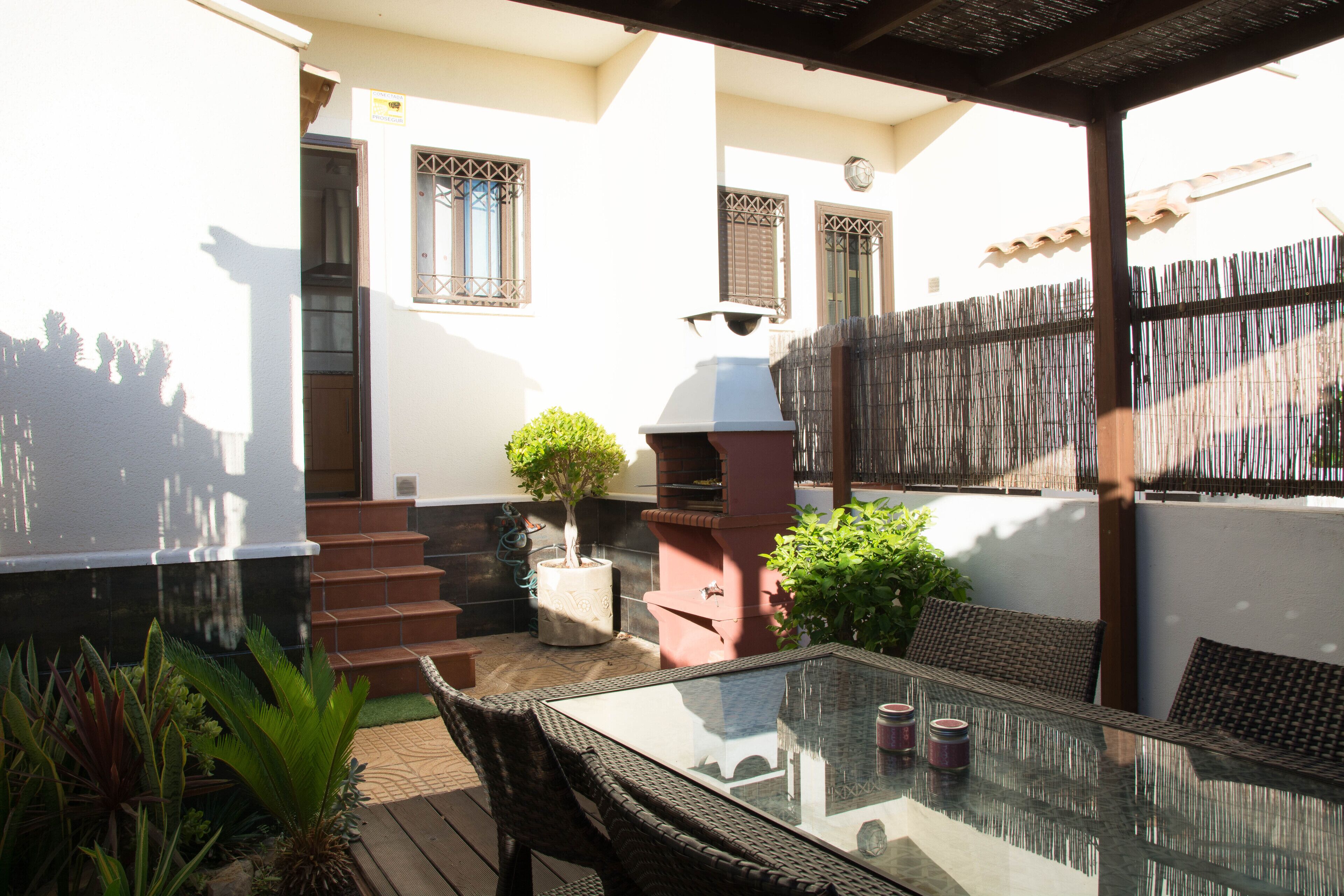 An outdoor patio with a glass table, wicker chairs, a barbecue grill, and a small tree.