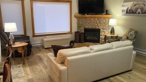 A living room with a stone fireplace, a flat-screen TV, a leather sofa, and a dining area with a table and chairs. - Boutique mountain condo with BREATH-TAKING views in the heart of it all! (Canmore)
