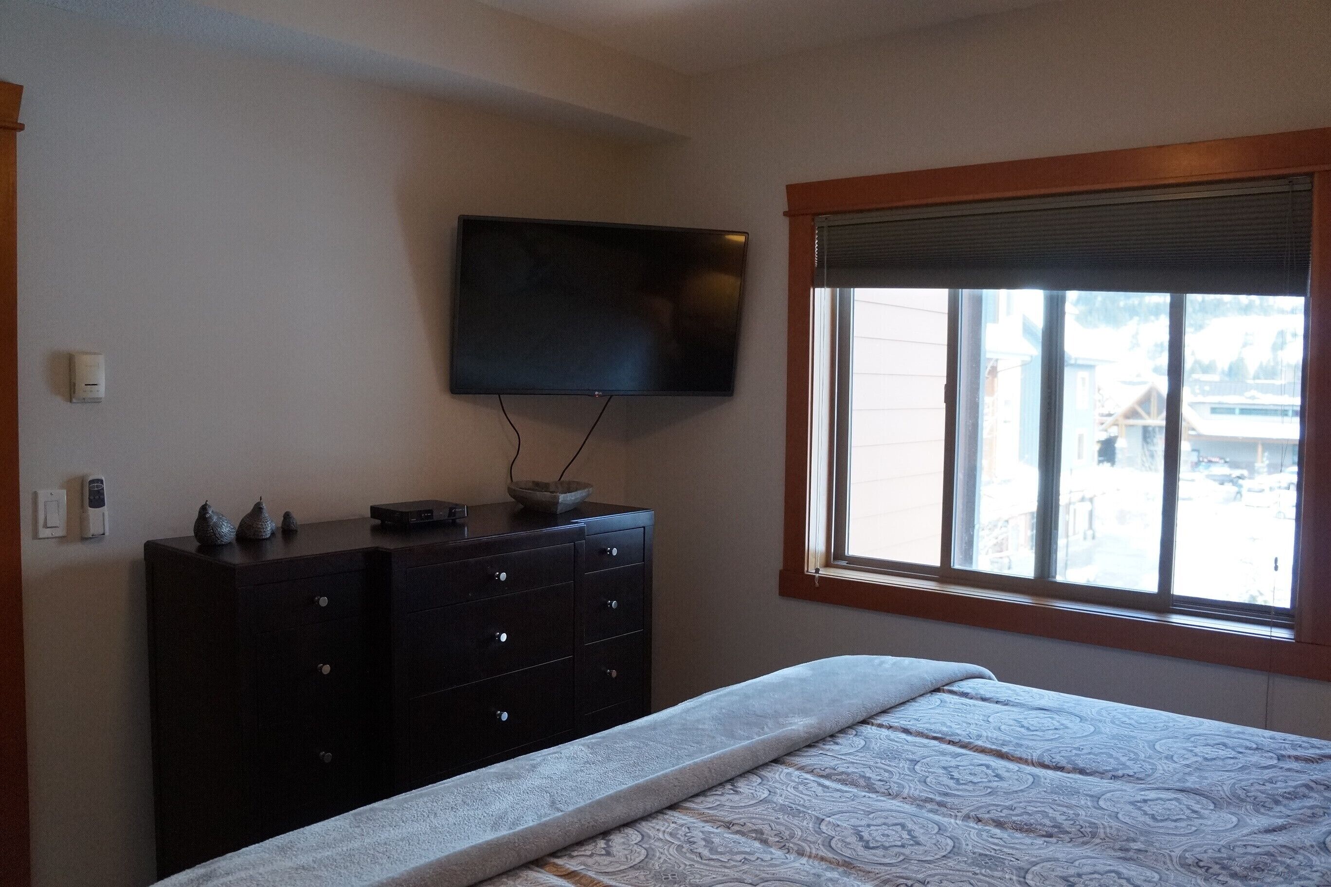 Quarto com cama, penteadeira, TV de tela plana na parede e janela com vista para prédios.