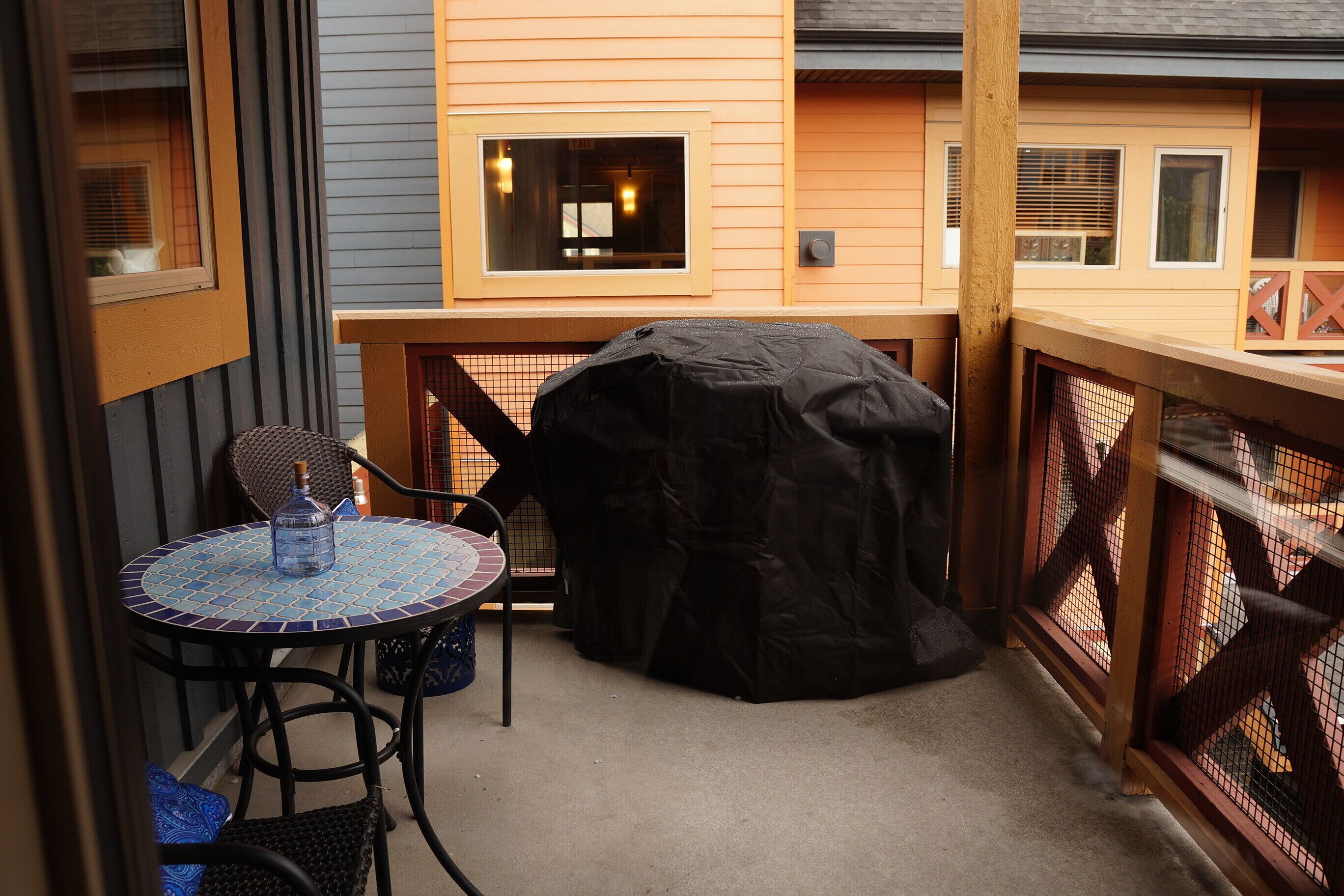 Une terrasse couverte avec une table et des chaises, un barbecue et une vue sur un immeuble doté de nombreuses fenêtres et d’un balcon.