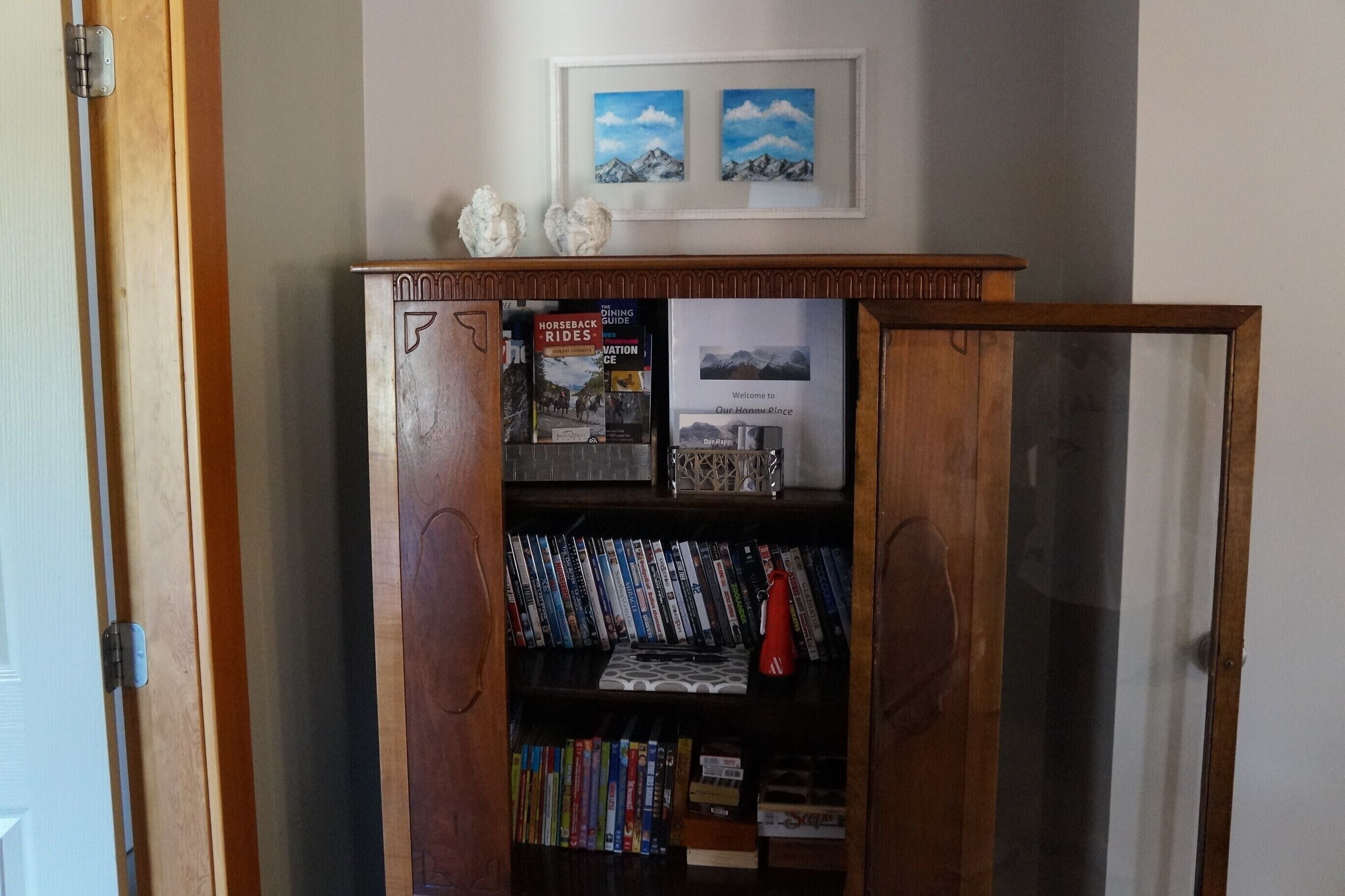 A wooden bookshelf with books and decorative items, a framed welcome sign, and two framed pictures on the wall.