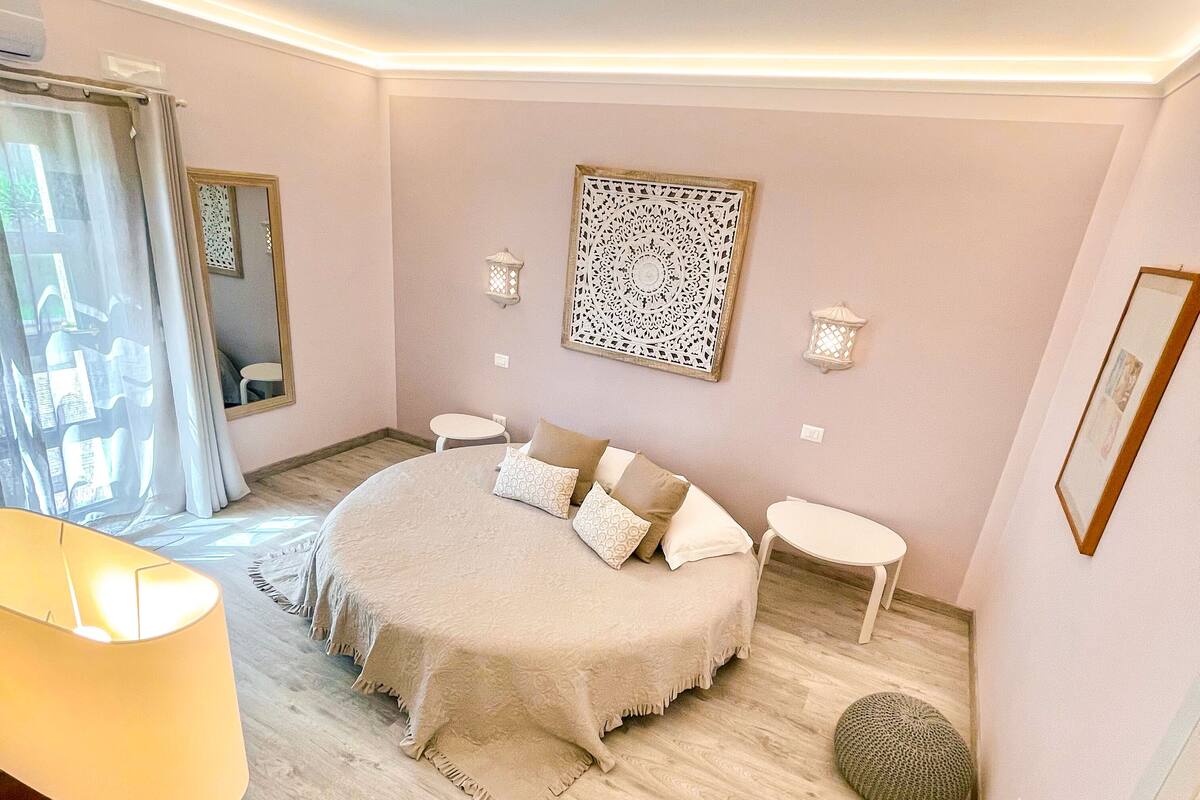 A bedroom with a round bed, bedside tables, a mirror, decorative wall art, and a window with curtains.