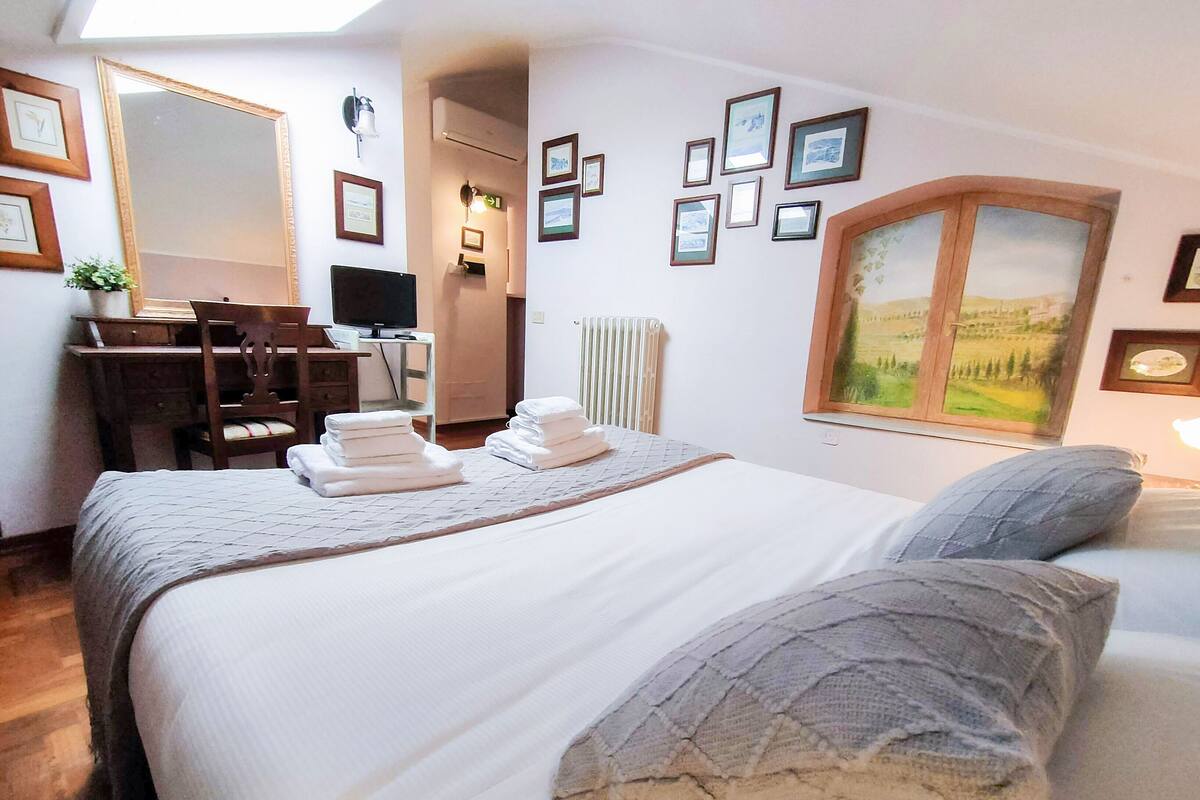 A neatly arranged bedroom with a bed, pillows, a desk with a chair, a television, and framed pictures on the wall.