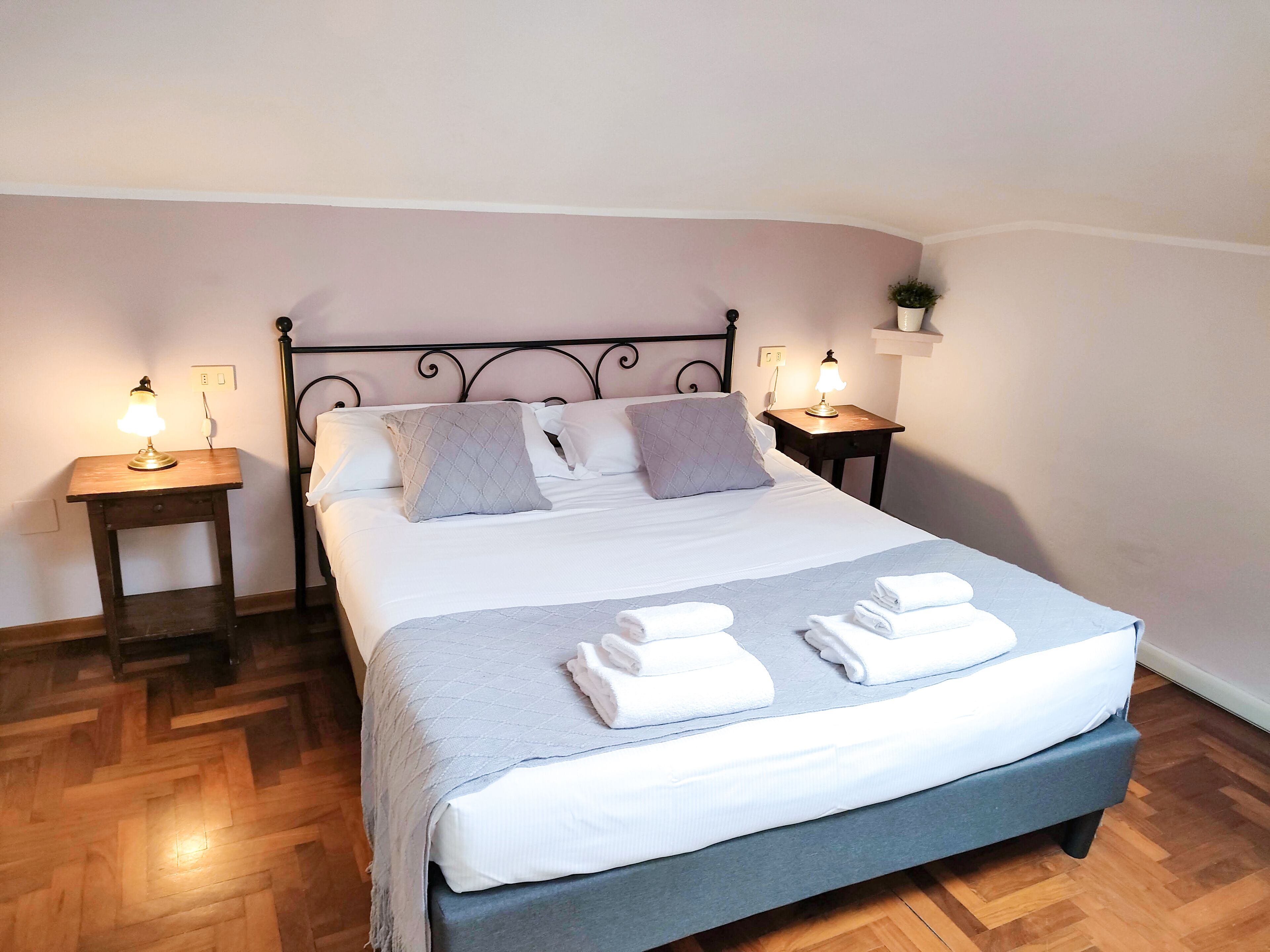 Een netjes opgemaakt bed met een grijze sprei en witte lakens, met aan weerszijden nachtkastjes met lampen, in een kamer met houten vloer en een eenvoudig, strak design.