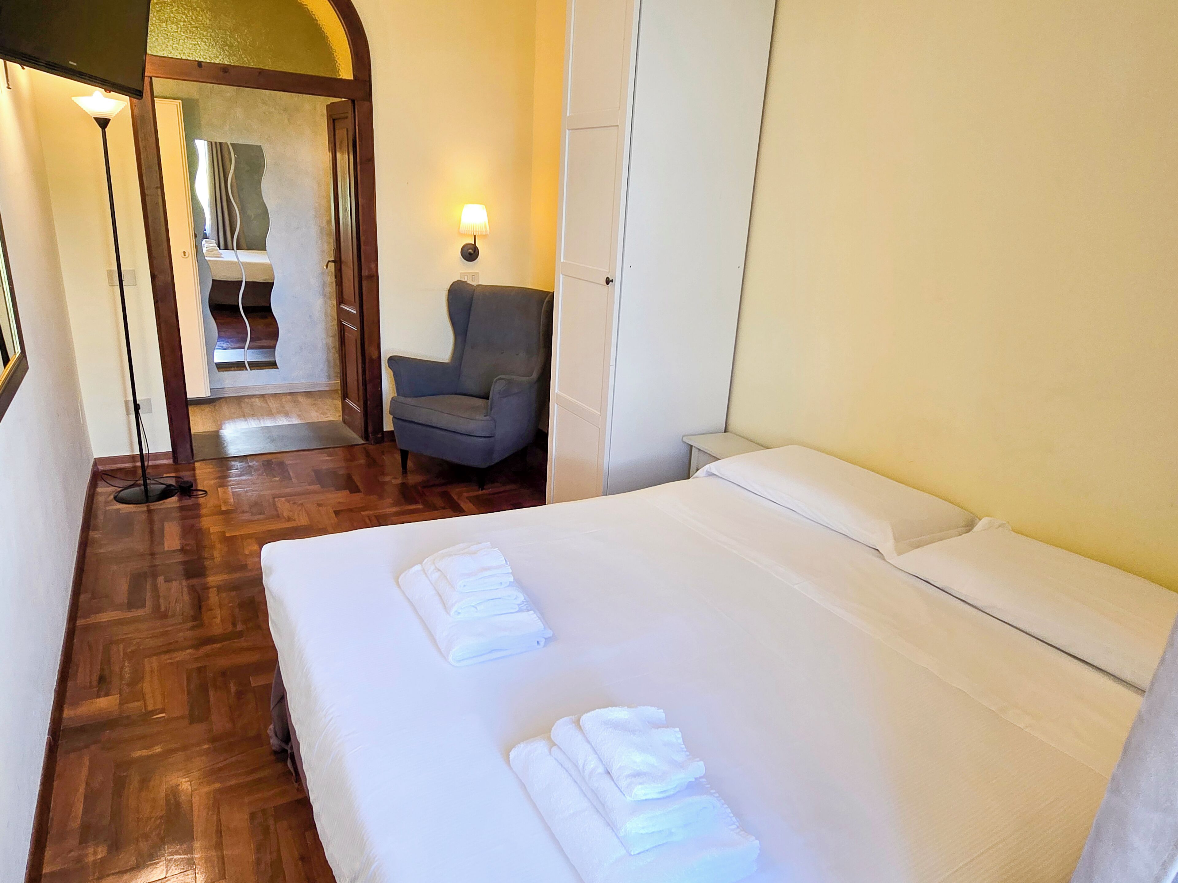 Une chambre d’hôtel avec un plancher en bois, un lit avec du linge de maison blanc, un fauteuil gris et une lampe.