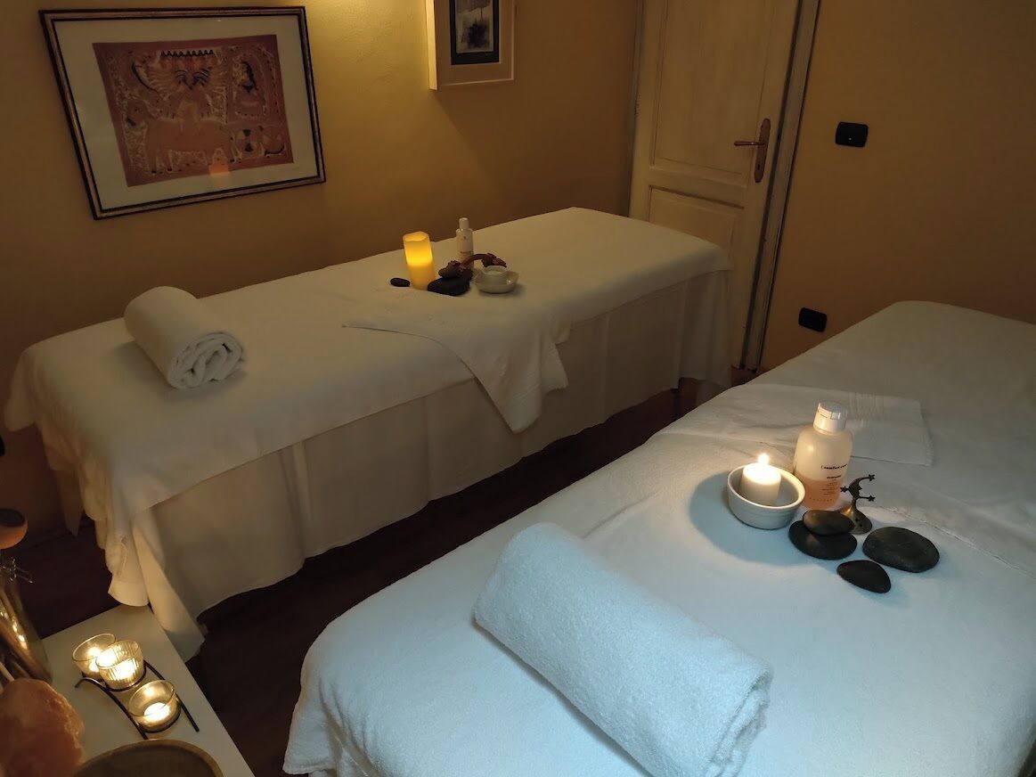 A room with two beds, each with a white bedspread and a rolled white towel. There are candles on the beds, a framed picture on the wall, and a small table with a candle and a bottle.