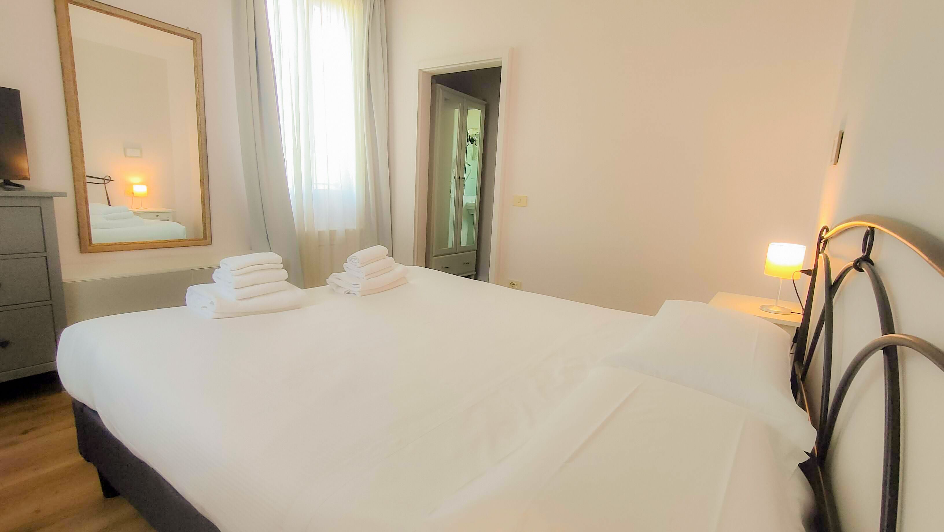 A neatly made bed with folded towels, a bedside lamp, a mirror, and a wardrobe in a hotel room.