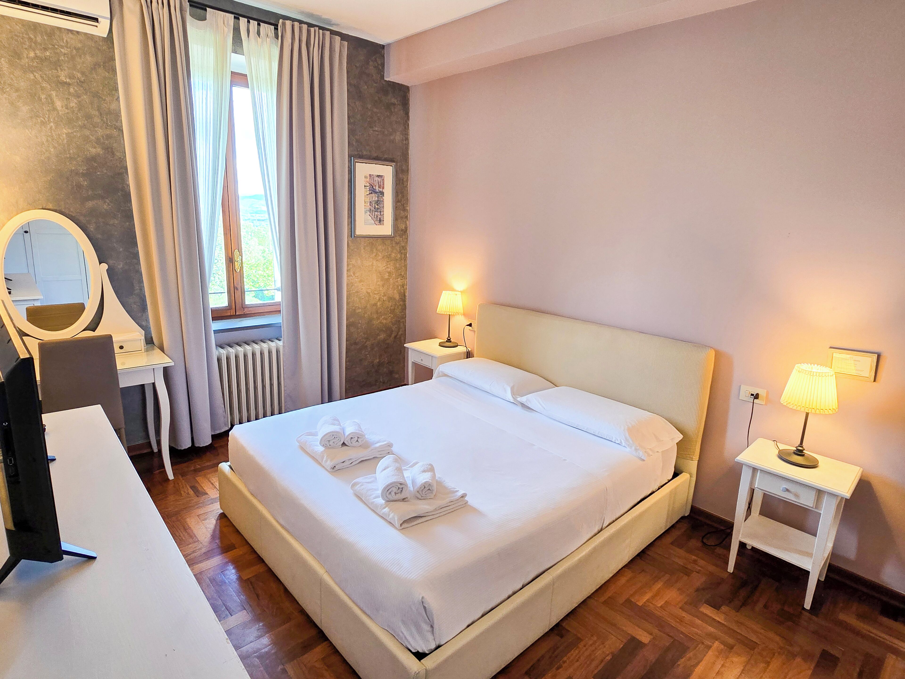 A neatly made bed with towels, a bedside lamp, a small table, a mirror, and a window with curtains.