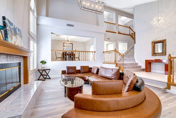 A spacious living room with a curved leather sofa, a glass coffee table, and a fireplace. A staircase with wooden railings leads to an upper level.