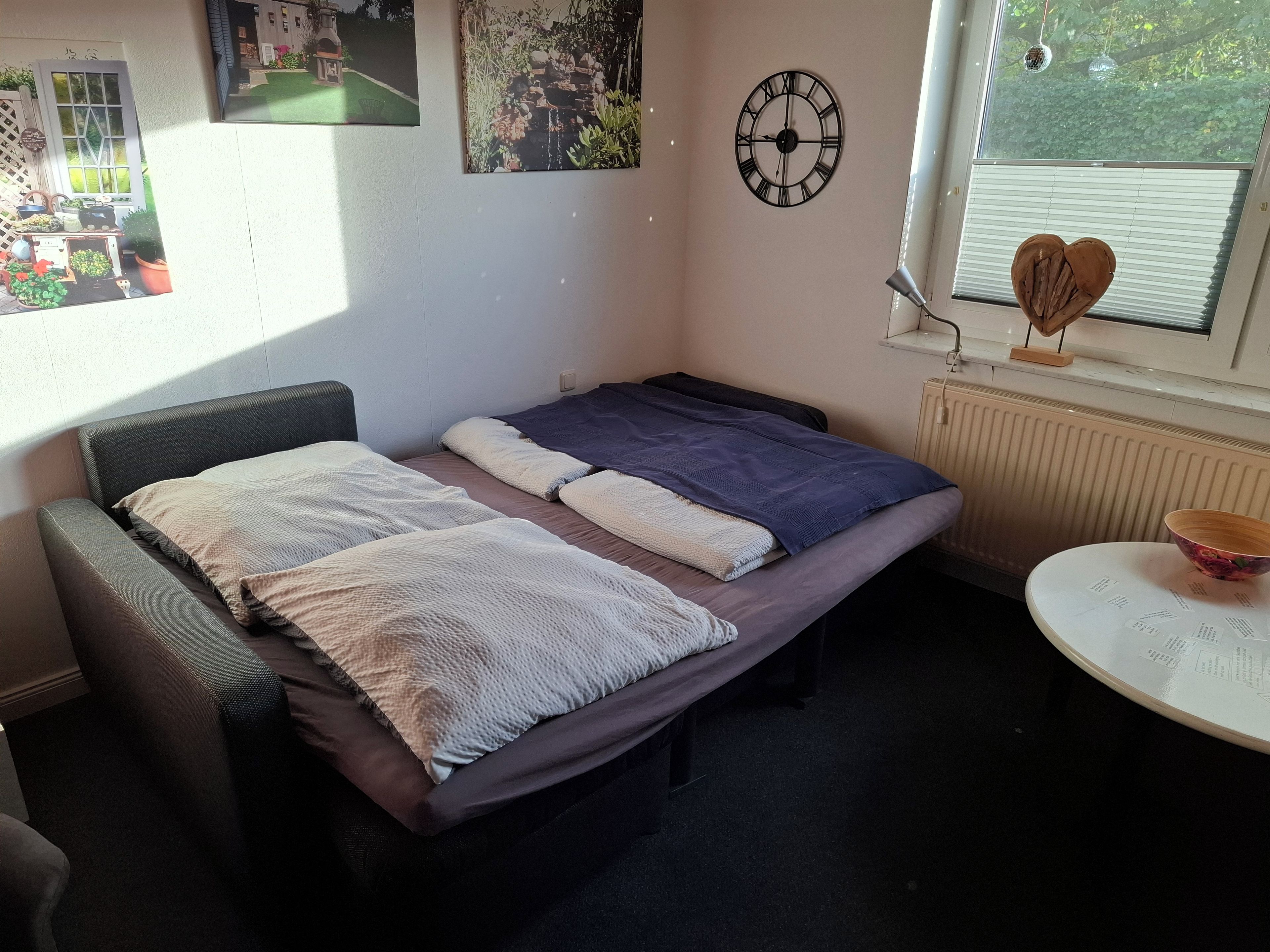 Una habitación con una cama, una mesita con un cuenco, un reloj y una ventana con persianas.