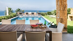 Un espace repas aménagé au bord de la piscine, avec vue sur l’océan, une table en bois et une pastèque posée sur la table.