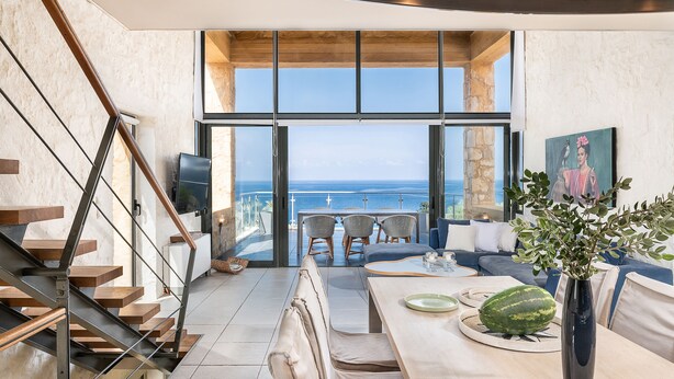 Un intérieur moderne avec un escalier, un coin repas et de grandes fenêtres offrant une vue sur la mer.