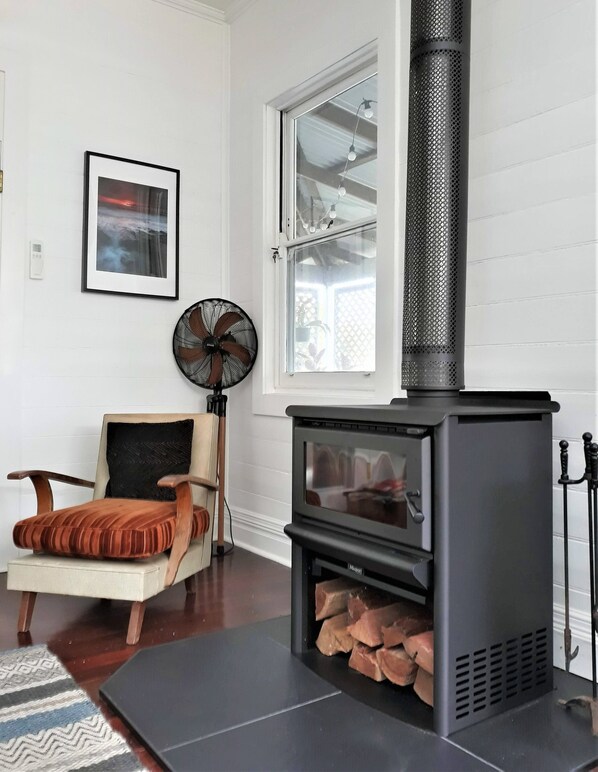 A modern living room with a wood-burning stove, a comfortable chair, a standing fan, and a framed picture on the wall. - Torodes - pet friendly and moments from the sand (Catherine Hill Bay)