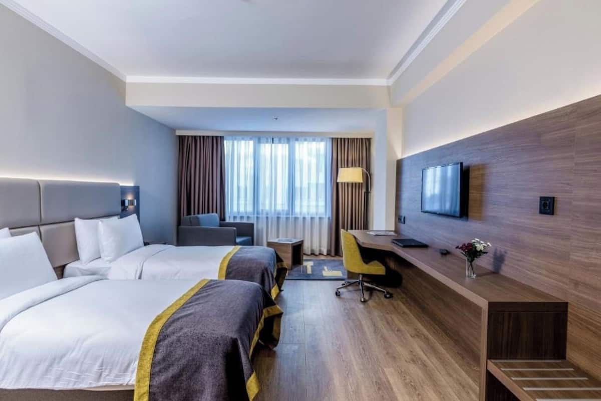A hotel room with two beds, a desk, a chair, a TV, and a window with curtains.
