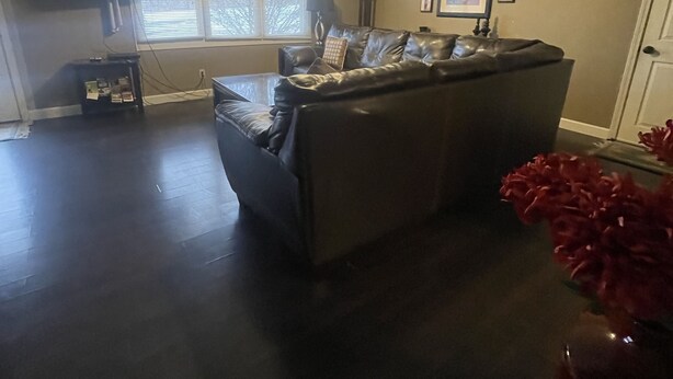 A living room with a dark hardwood floor, a leather sofa, a ceiling fan, and a flat-screen TV mounted on the wall.