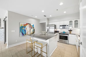 Una cocina moderna con armarios blancos, electrodomésticos de acero inoxidable, una isla central con taburetes altos y un cuadro abstracto colorido en la pared.