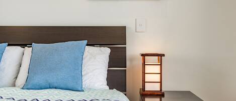 A neatly made bed with a patterned bedspread, a dark headboard, and two pillows. A small bedside table with a lamp is visible to the right.