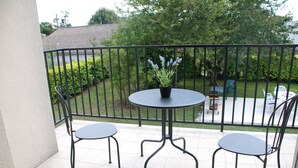 A balcony with a round table and two chairs, a potted plant, and a view of a garden and street.