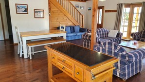 Un salon chaleureux avec une table à manger en bois, des canapés bleus et à carreaux, un escalier et une grande fenêtre avec des rideaux.