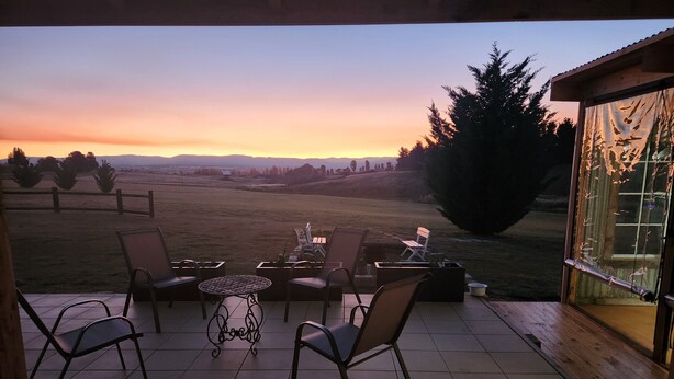 Une terrasse avec des sièges d’extérieur, une petite table et une vue sur un coucher de soleil se reflétant sur un champ verdoyant.