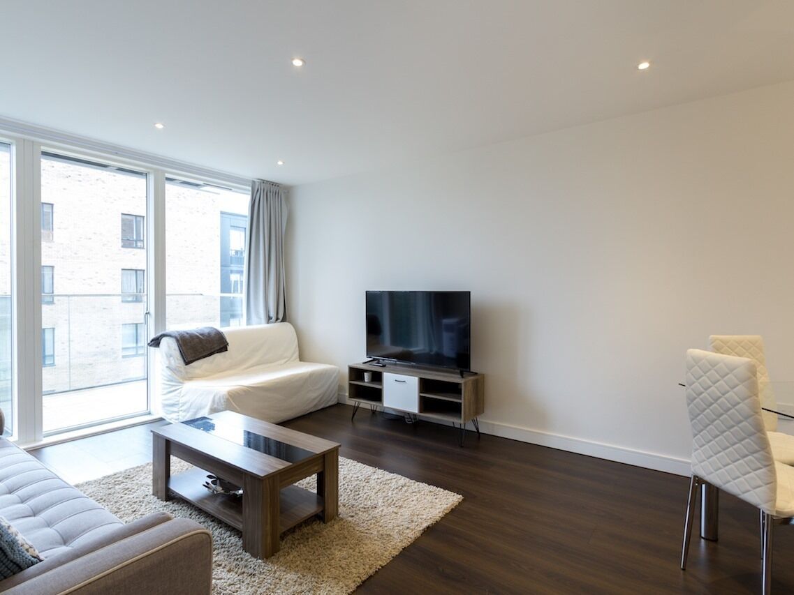 A modern living room with a sofa, a coffee table, a TV on a stand, and a view of the outside through large windows.