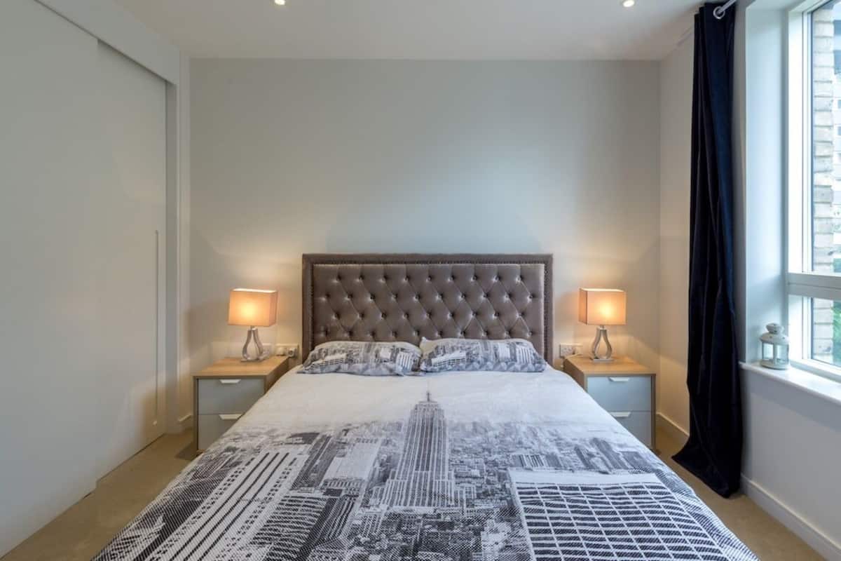 Un dormitorio con cabecera capitonada, cama con edredón de diseño urbano, dos lámparas de noche y una ventana con cortina.