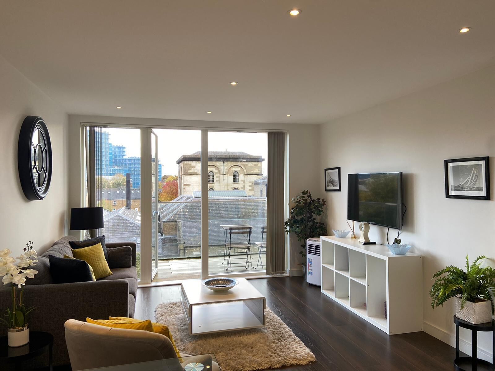 A modern living room with a sofa, coffee table, and TV stand. There is a large window with a view of buildings outside.
