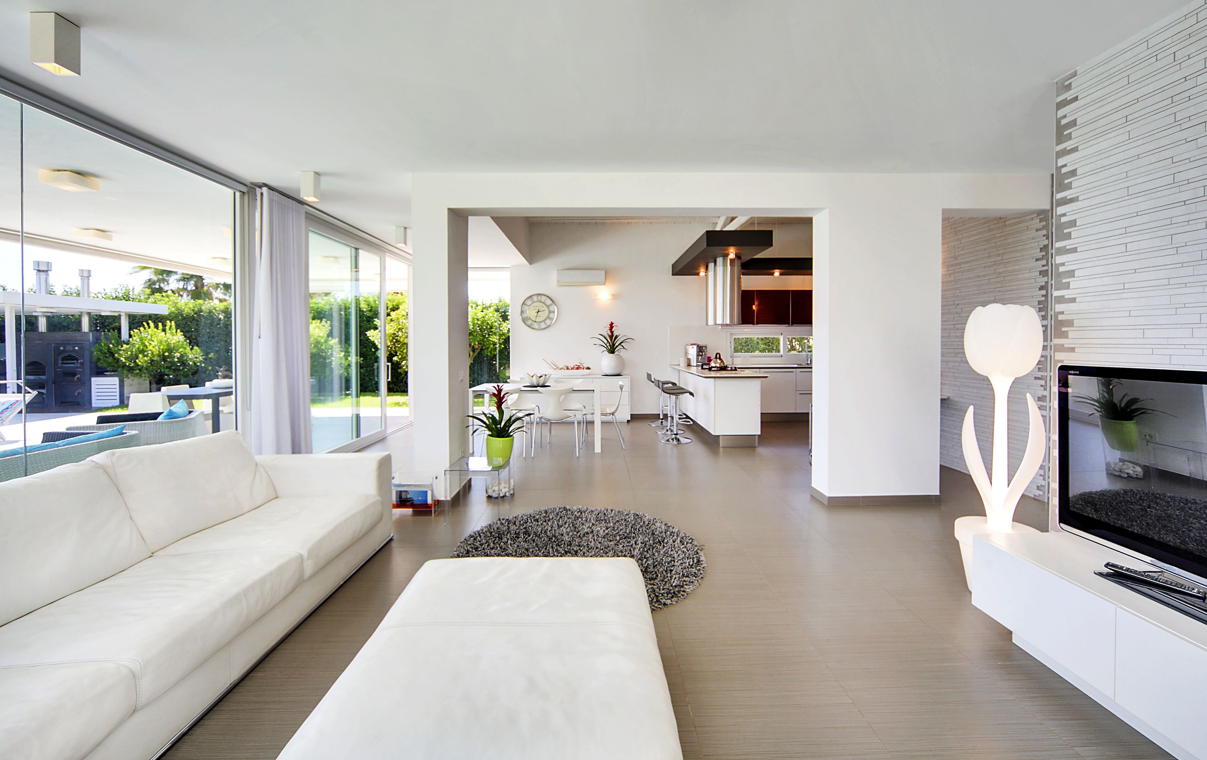 A modern living room with a white sofa, a dining area with a white table and chairs, and a kitchen with white cabinets and a large island.