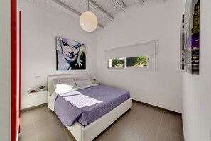 A modern bedroom with a large bed, a painting on the wall, and a window with a view of greenery.