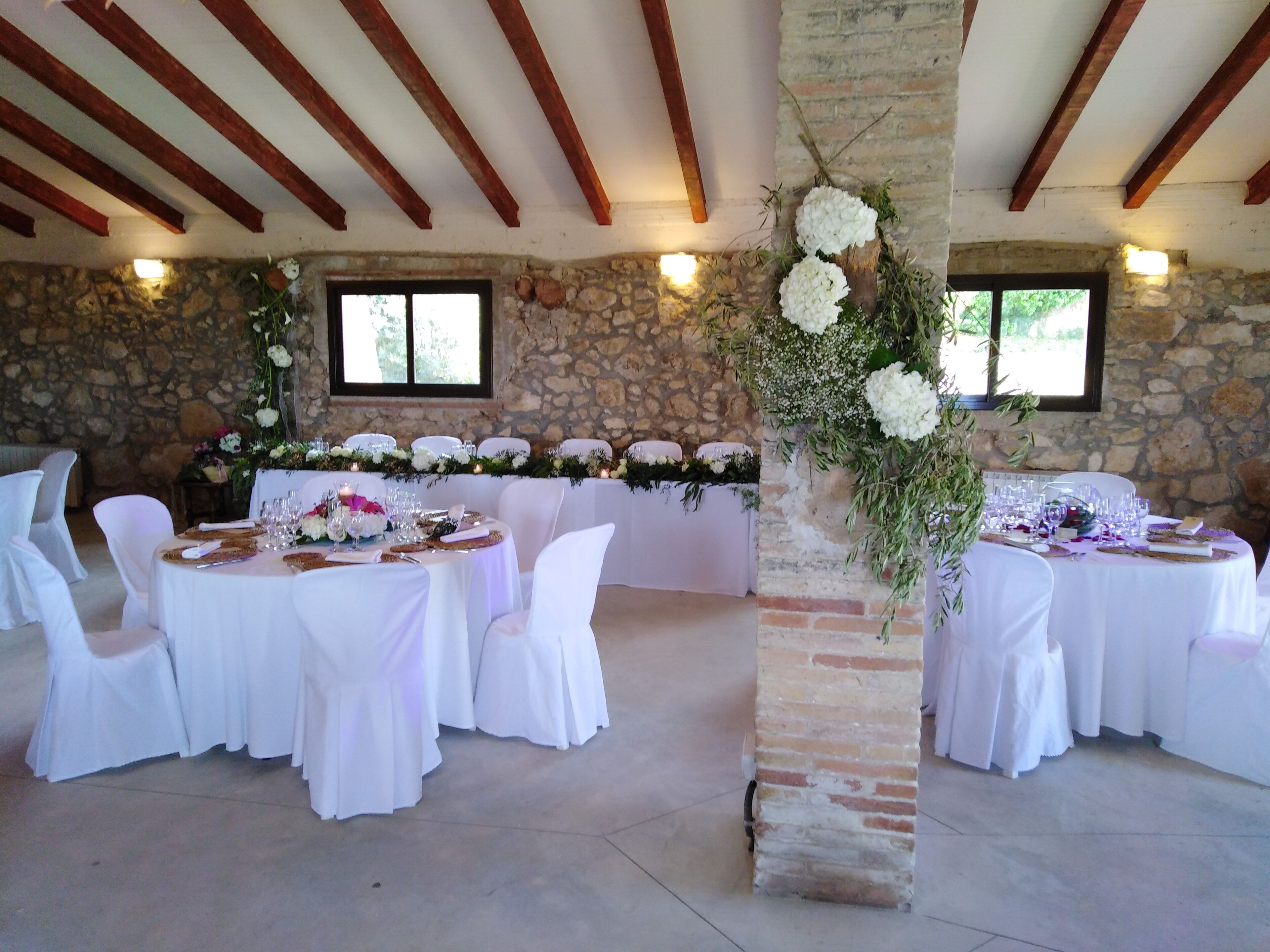 Un salón de banquetes con mesas redondas, sillas blancas, paredes de piedra y vigas de madera.