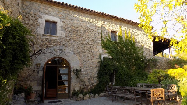 A stone building with a wooden door and windows, surrounded by greenery and a bench.