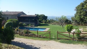 A rustic stone building with a swimming pool, surrounded by a wooden fence and lush greenery.