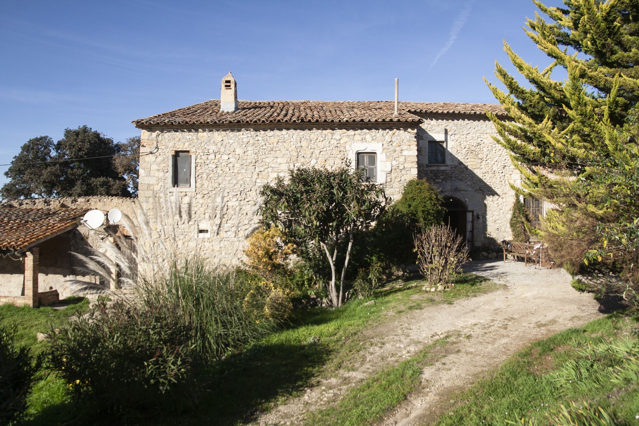 A stone house with a tiled roof, a dirt path leading to the entrance, and surrounding vegetation.
