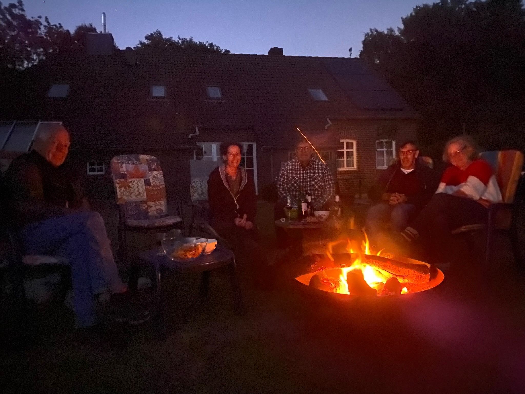 Un grupo de personas sentadas alrededor de una fogata en un entorno al aire libre, con una casa al fondo.