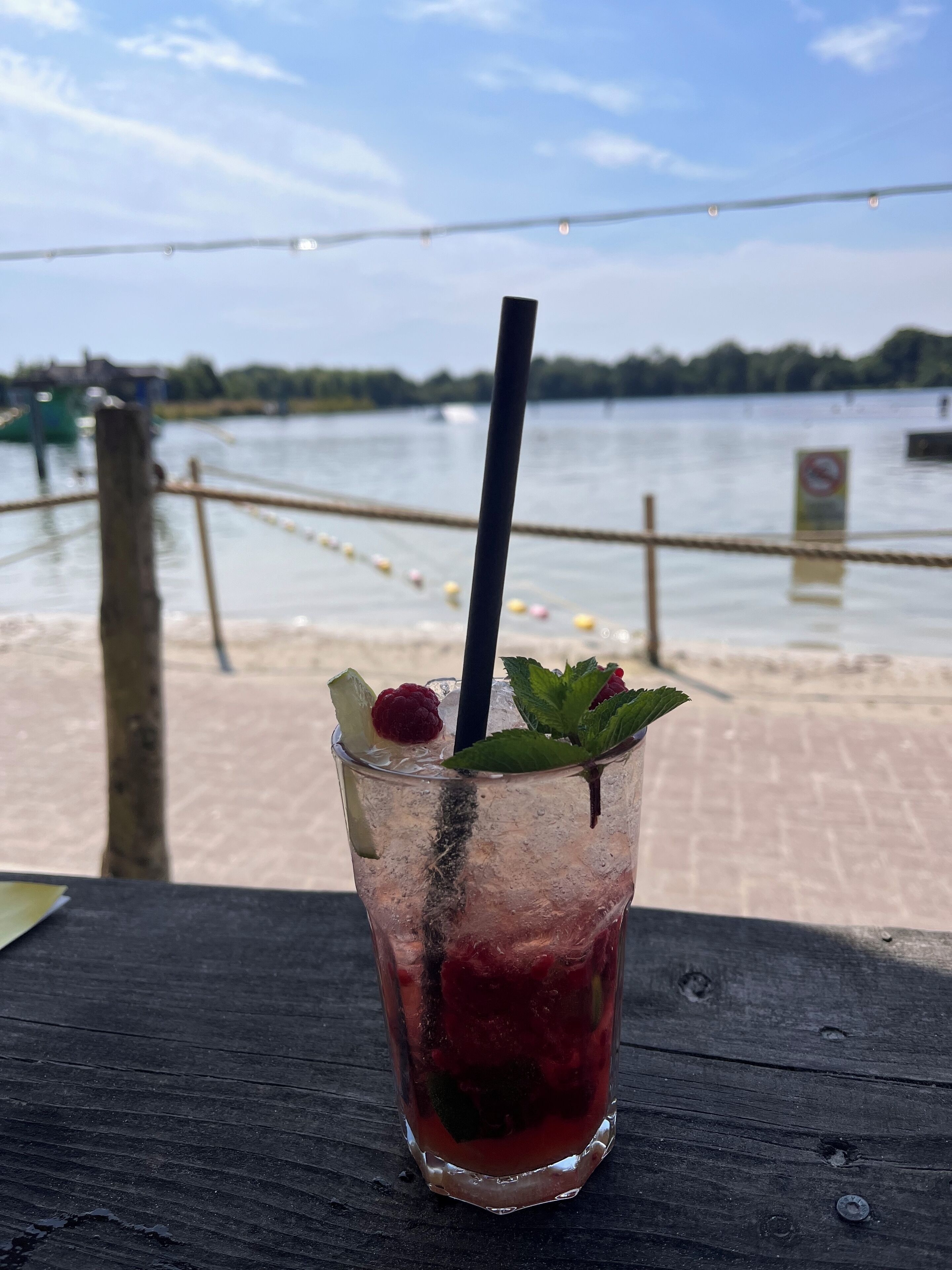 A glass of iced drink with a straw and mint garnish on a wooden table by a waterfront.