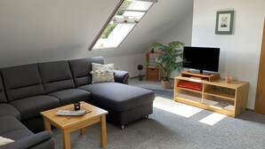 A modern living room with a sectional sofa, a small wooden table, a TV on a wooden stand, and a skylight. - Idyllically situated. 90m2 apartment for families + travelers up to 5 persons + garden + terrace near the North Sea 🐟 (Marienhafen)