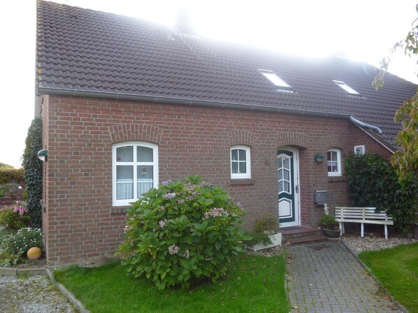 A brick house with a tiled roof, a front door, and a small garden with a bench and potted plants. - Idyllically situated. 90m2 apartment for families + travelers up to 5 persons + garden + terrace near the North Sea 🐟 (Marienhafen)