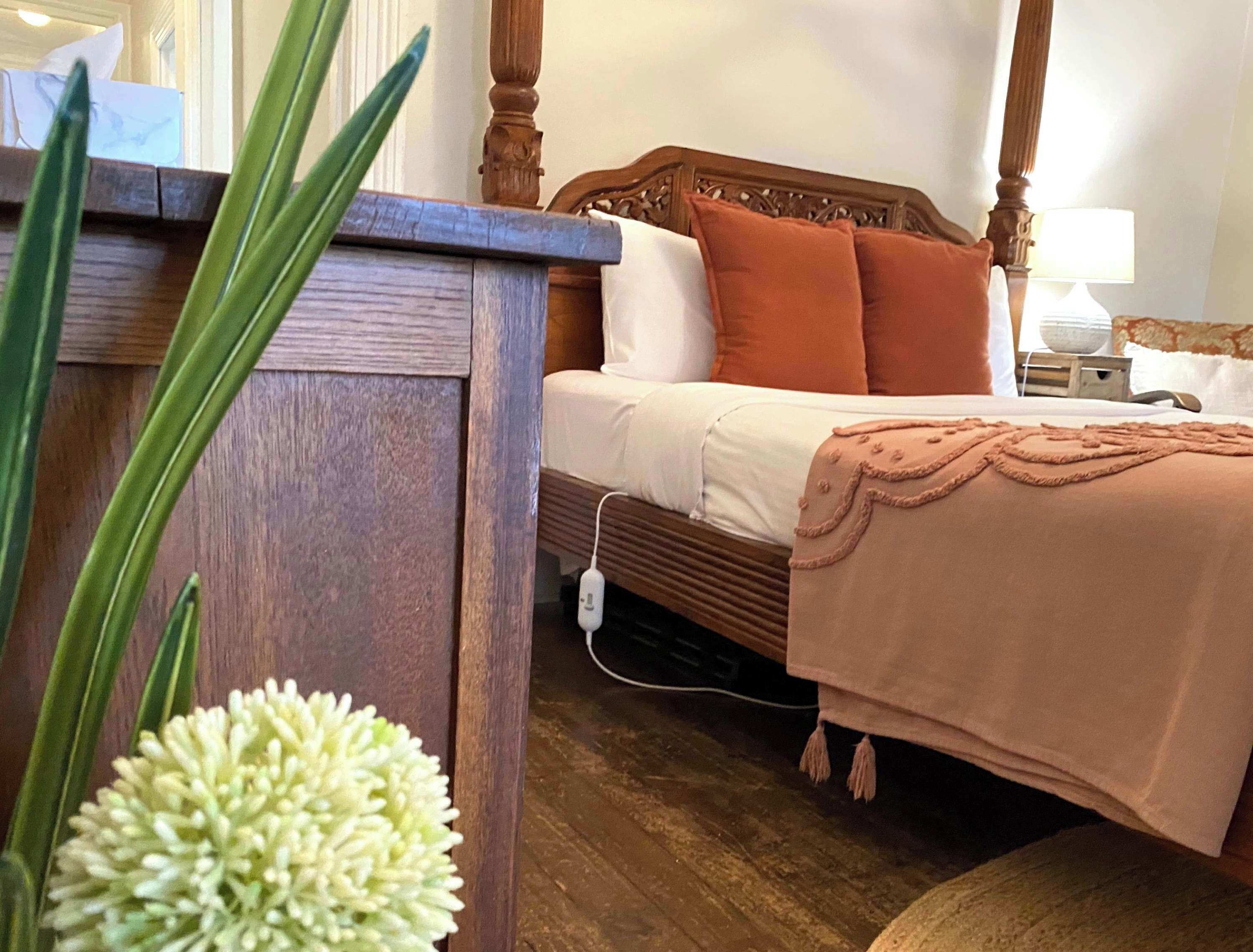 A wooden bed with white and orange pillows, a bedside table with a power outlet, and a white flower vase.
