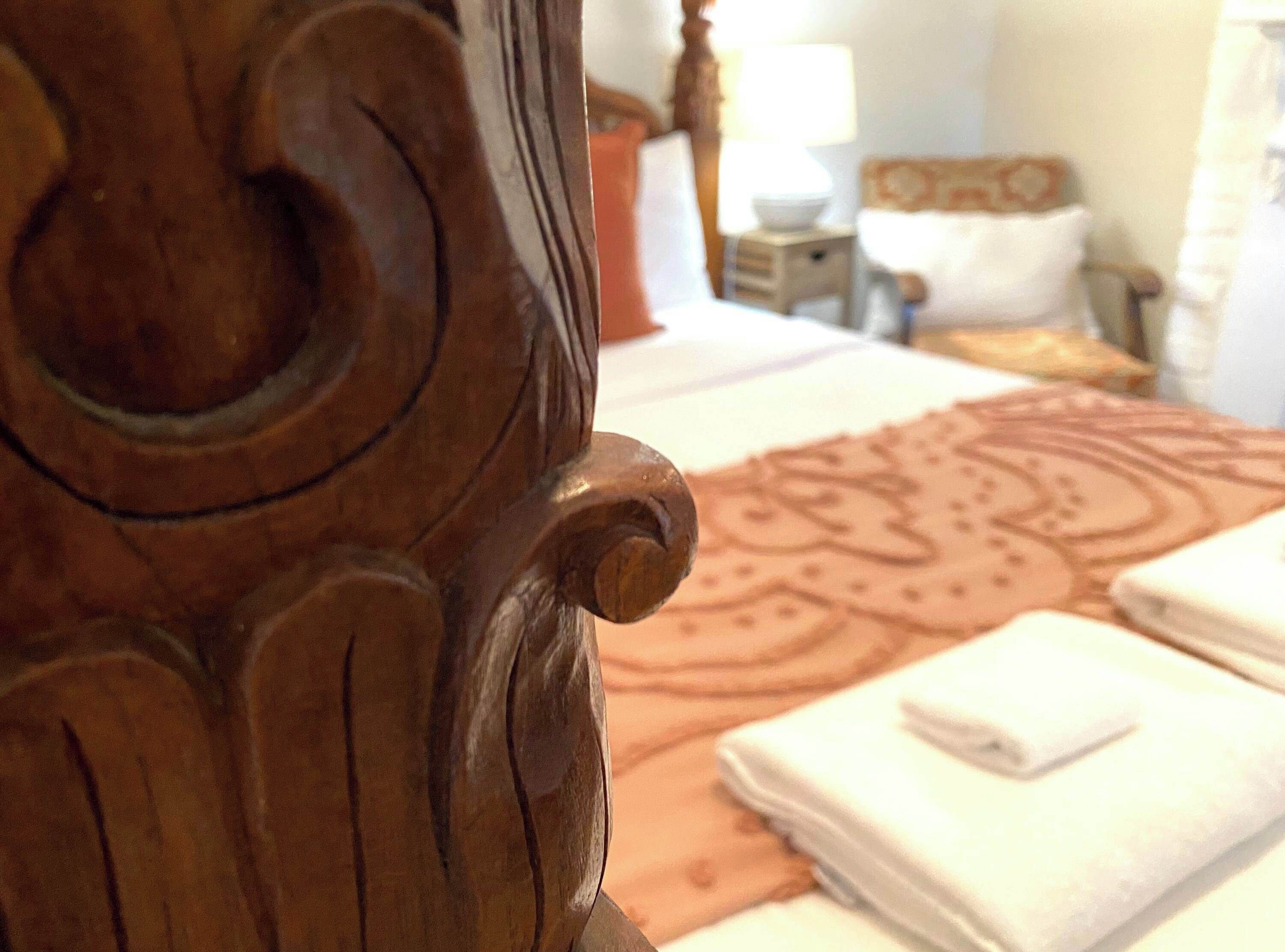 A close-up of a wooden bed headboard with carved details, a patterned bedspread, and neatly folded white towels on a bed.