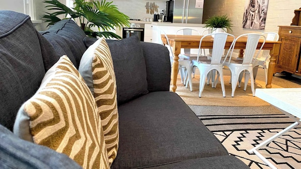 Uma sala de estar moderna com um sofá cinzento, almofadas estampadas, uma área de refeições com cadeiras brancas, uma mesa de madeira e uma cozinha ao fundo.