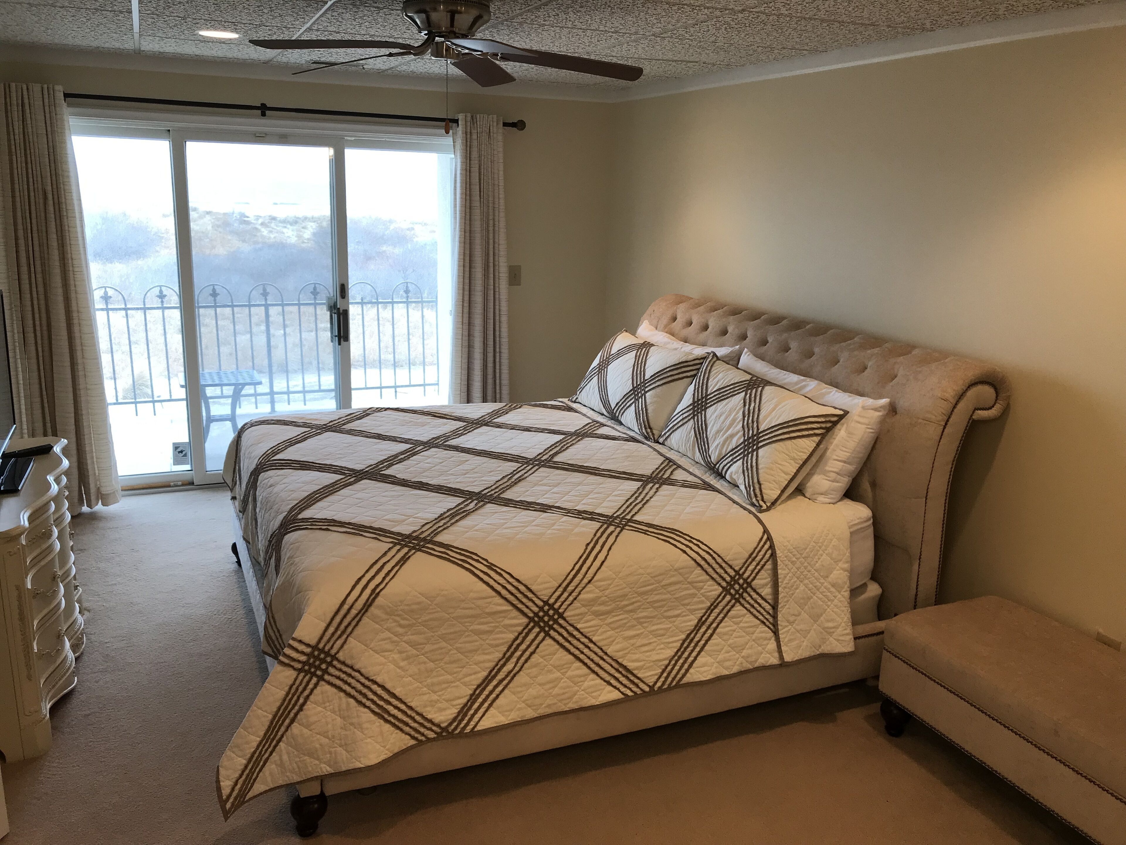 A bedroom with a large bed, a ceiling fan, a sliding glass door to a balcony, and a bedside table.