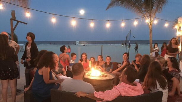 A group of people gathered around a fire pit on a beach at dusk, with string lights overhead.